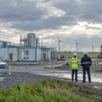 Wasserstoffanlage mit Rohrleitungen, Tanks und Containern; zwei Arbeiter mit Schutzhelm auf einer schlammigen Baustelle, dahinter ein Kran und Windräder unter bewölktem Himmel.
